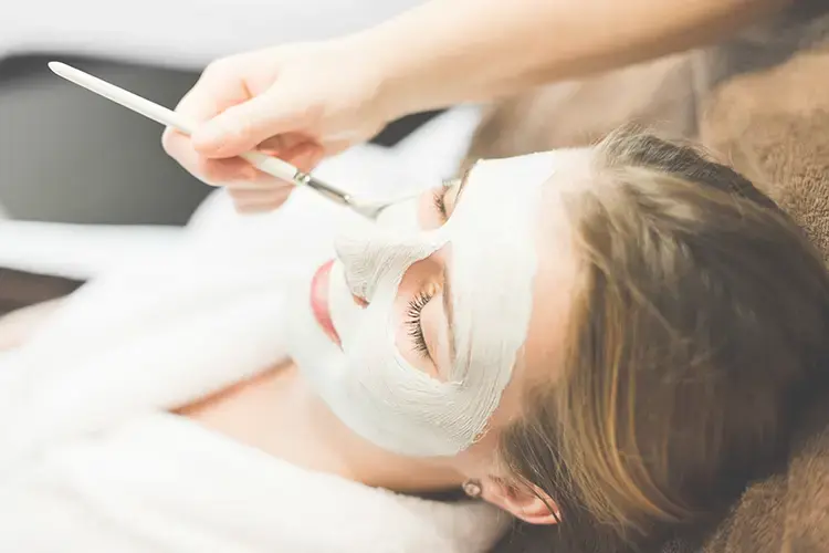 Frau im Bademantel auf einer Liege. Mit einem Pinsel wird eine Gesichtsmaske aufgetragen.