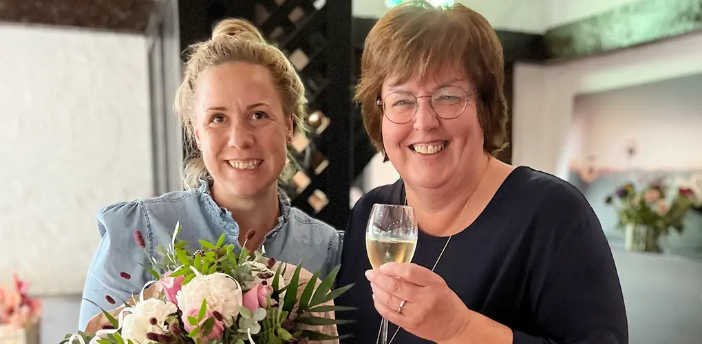 Hoteldirektorin Jennifer Kürsten und Ute Lenarz posieren mit Sektglas und Blumenstrauß