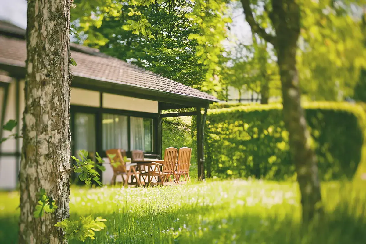 Blick über eine Grüne Wiese durch Bäume hindurch auf eine sonnige Terrasse eines Ferienhauses