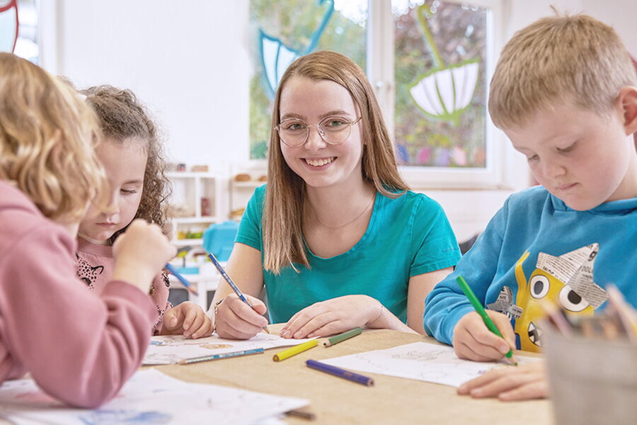 Kids Club Teamerin malt mit Kindern schöne Bilder und lächelt in die Kamera