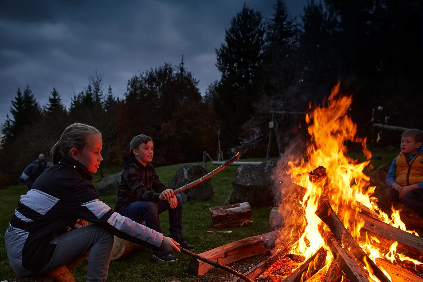 Kinder sitzen am Lagerfeuer und grillen Stockbrot bei Abenddämmerung Kinder sitzen am Lagerfeuer und grillen Stockbrot bei Abenddämmerung